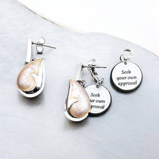 Silver nautilus shell earrings with charm tags that read 'Seek your own approval', beautifully arranged on a smooth grey surface - elegant, ocean-inspired, and uplifting.