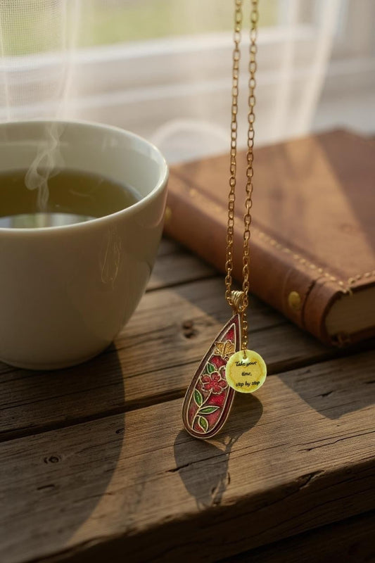 Floral enamel pendant necklace on a gold chain with a charm that reads 'Take your time, step by step', displayed on a wooden table between a cup of herbal tea and a leather-bound book.