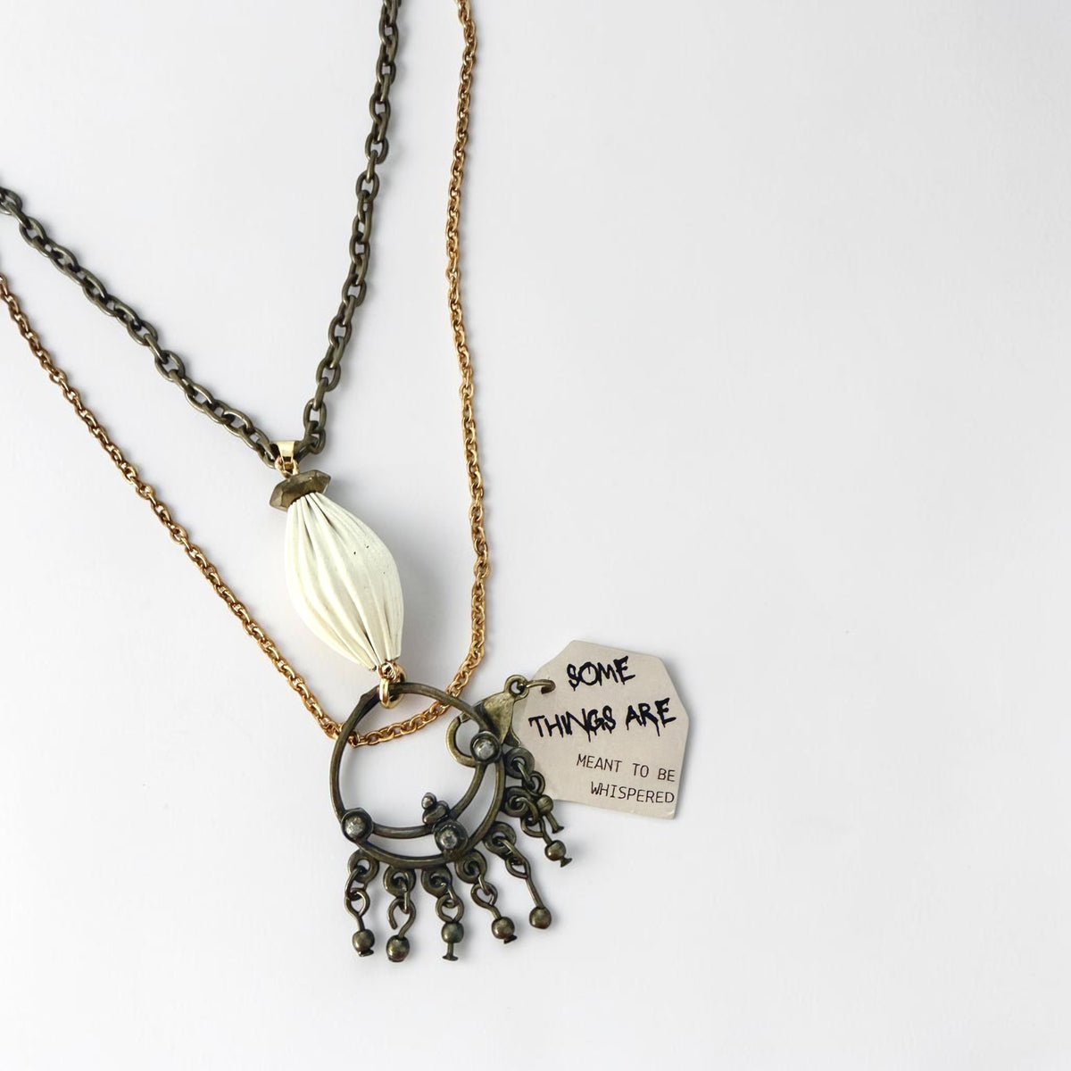 A close up of a necklace featuring an ivory lantern-inspired pendant, ornate antique bronze drop and reminder tag engraved with 'Some things are meant to be whispered.