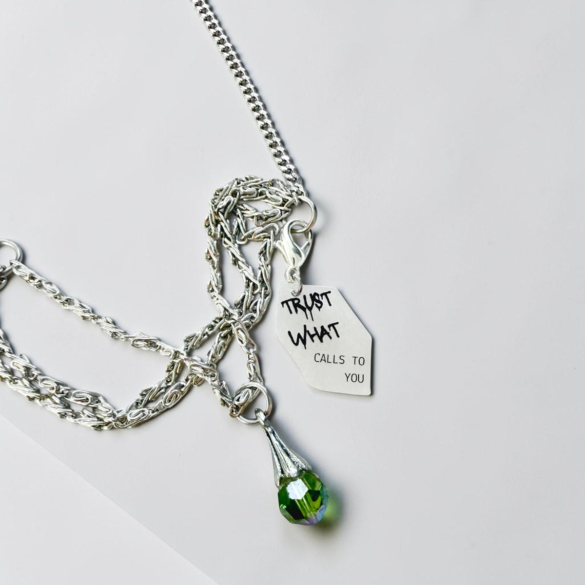 A silver-toned chain necklace with a small hanging tag that reads 'TRUST WHAT CALLS TO YOU' and a faceted green teardrop crystal pendant, photographed on a light grey background.