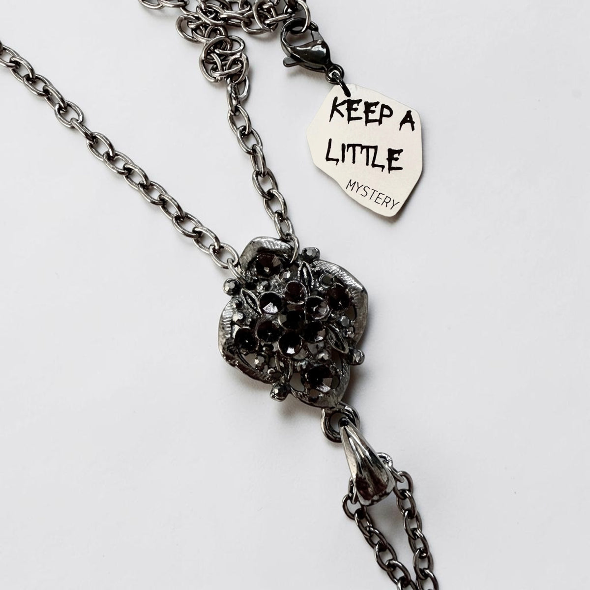 A dark gunmetal-toned chain necklace with an ornate pendant featuring a black faceted central stone, shown on a plain white background with an attached tag that reads 'KEEP A LITTLE MYSTERY'.