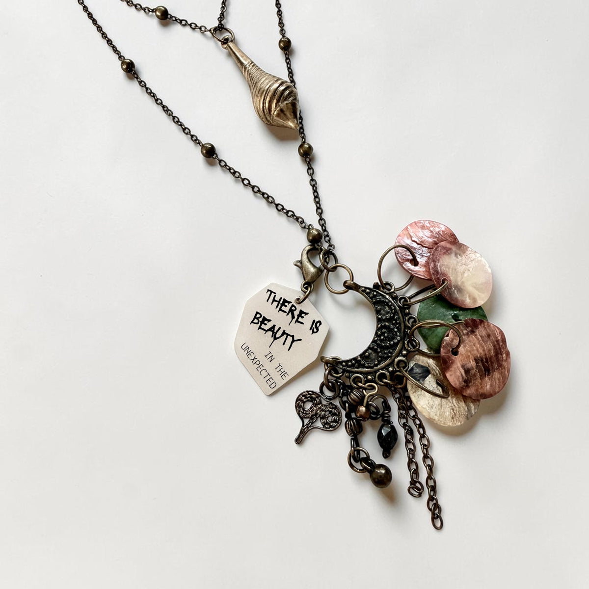 A bohemian-style layered necklace featuring a dark bronze chain with a spiral pendant, a tag engraved with 'THERE IS BEAUTY IN THE UNEXPECTED', a dark metal crescent moon with a metal heart charm, and multiple natural shell disc charms in pink, green, and beige tones.