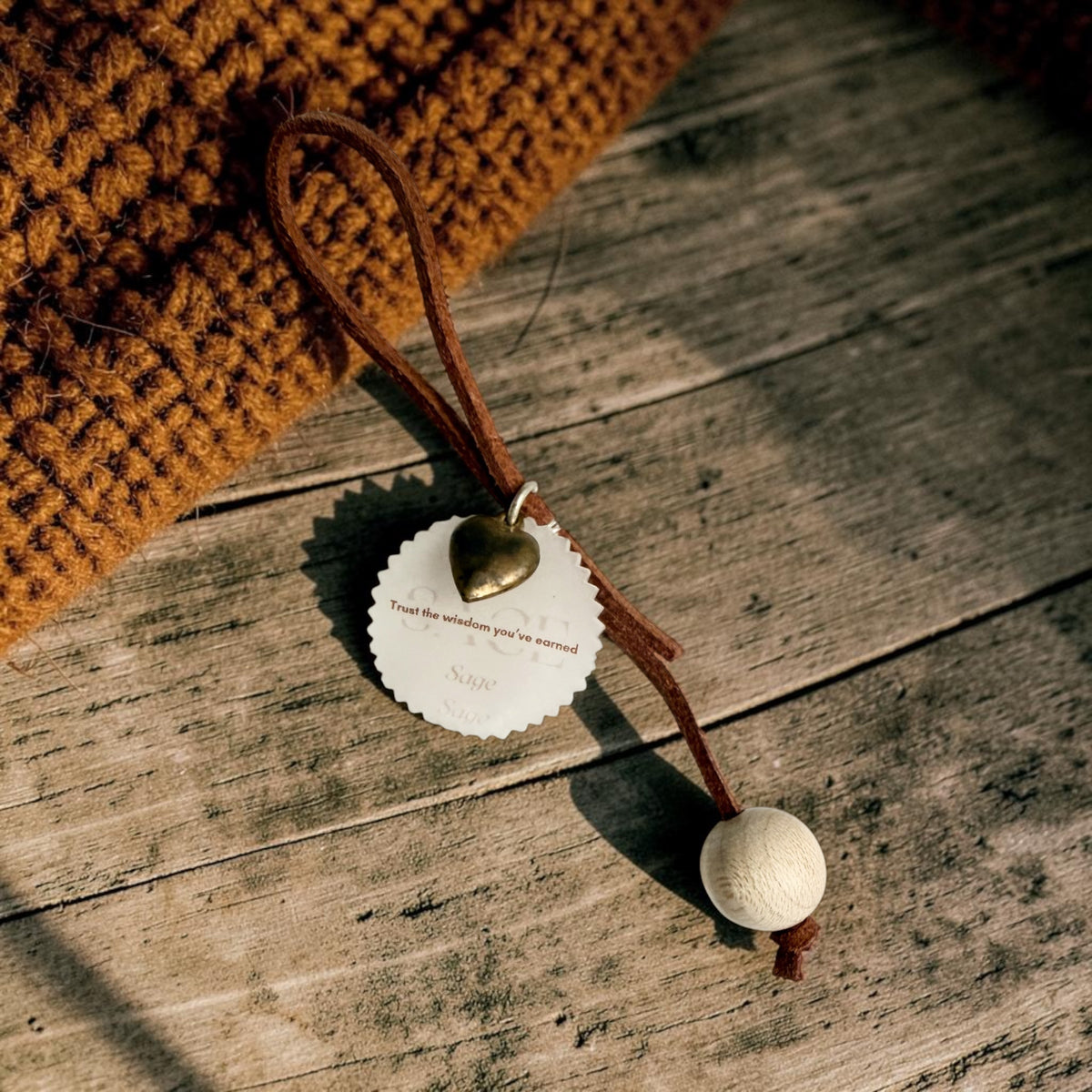 A brown cord ornament with a small brass heart, a round white tag, and a wooden bead, laid on a rustic wooden surface next to a chunky mustard knit fabric.