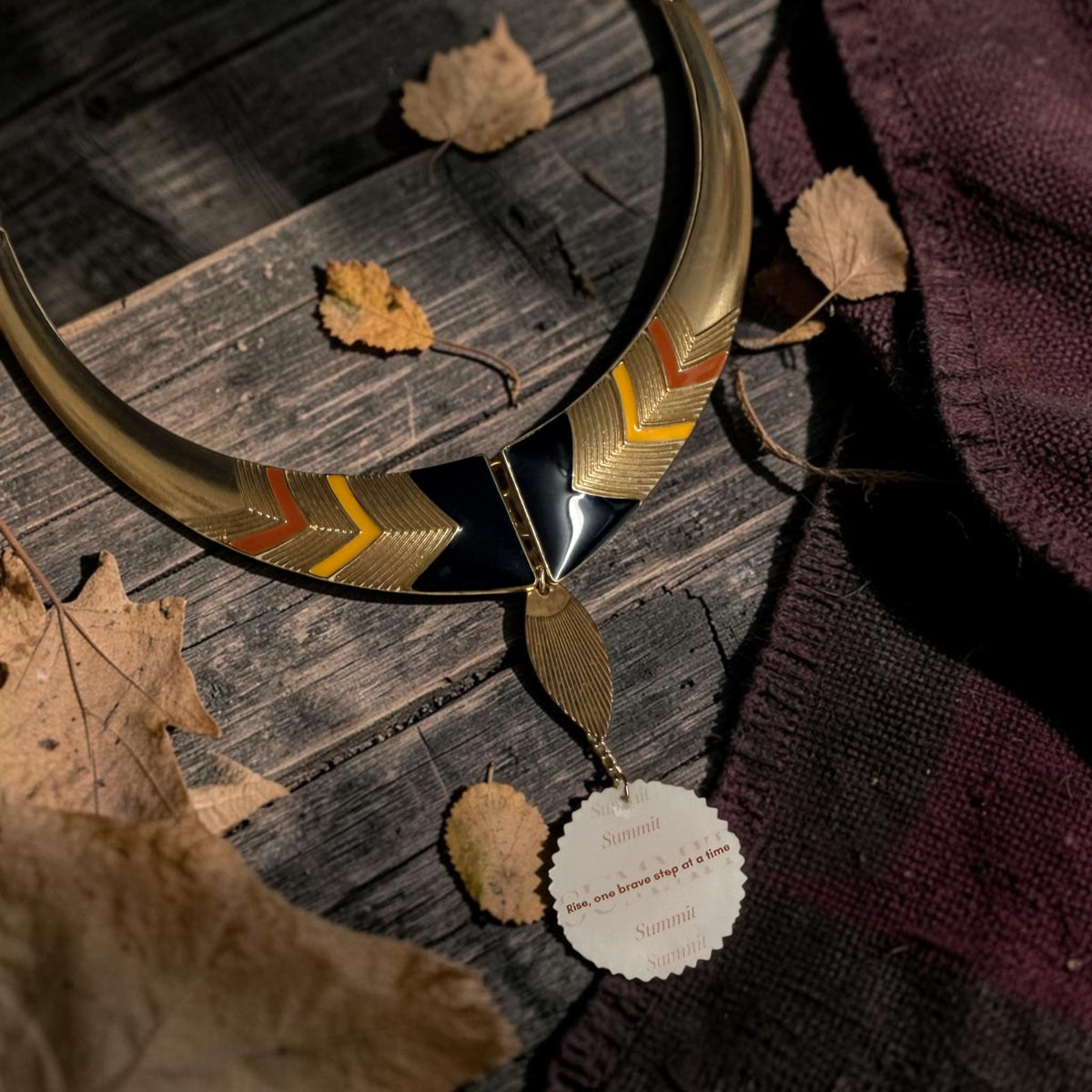 A gold-toned crescent collar necklace with geometric chevron patterns in black, orange, and yellow, displayed on a rustic wooden surface with dried leaves and a dark knit fabric.