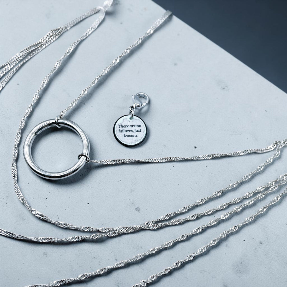 Layered stainless steel necklace with a large hoop pendant and an engraved tag reading ' There are no failures, just lessons', displayed on a grey textured background.