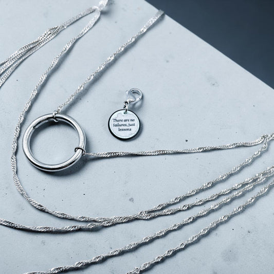 Layered stainless steel necklace with a large hoop pendant and an engraved tag reading ' There are no failures, just lessons', displayed on a grey textured background.
