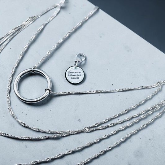 Layered stainless steel necklace with a large hoop pendant and an engraved tag reading ' There are no failures, just lessons', displayed on a grey textured background.