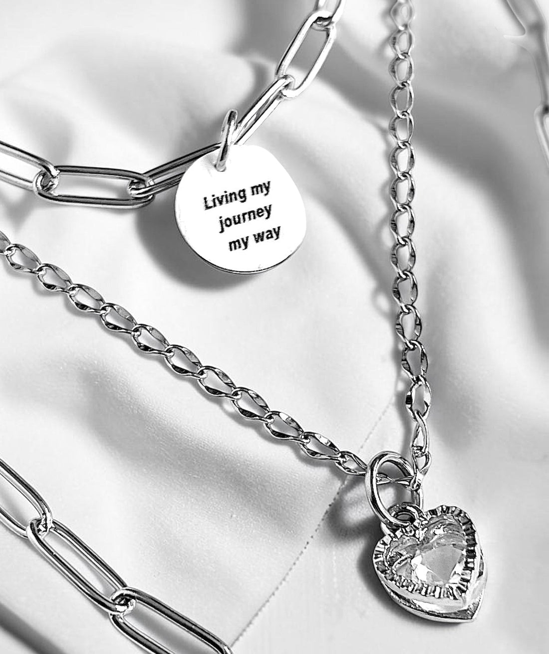 Layered stainless steel necklace on white fabric featuring an engraved tag charm reading 'Living my Journey My Way' with a sparkling crystal heart pendant.