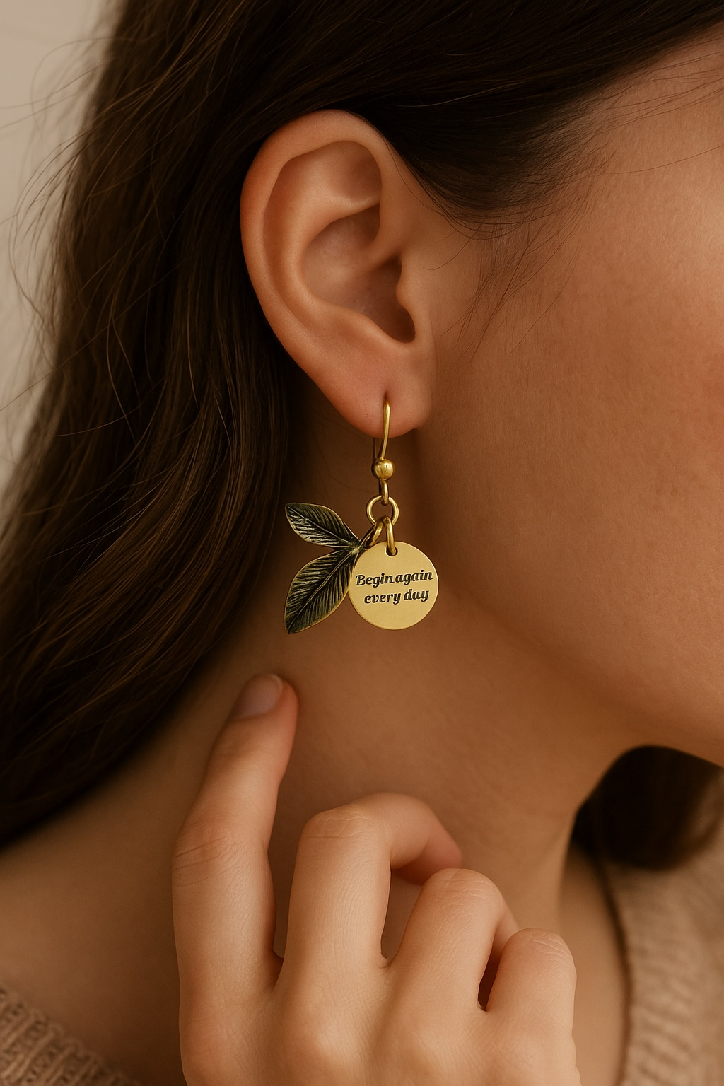 A close-up of a woman wearing an antique bronze/gold leaf-shaped earring. Attached to the earring is a small charm tag that reads, 'Begin again every day.'