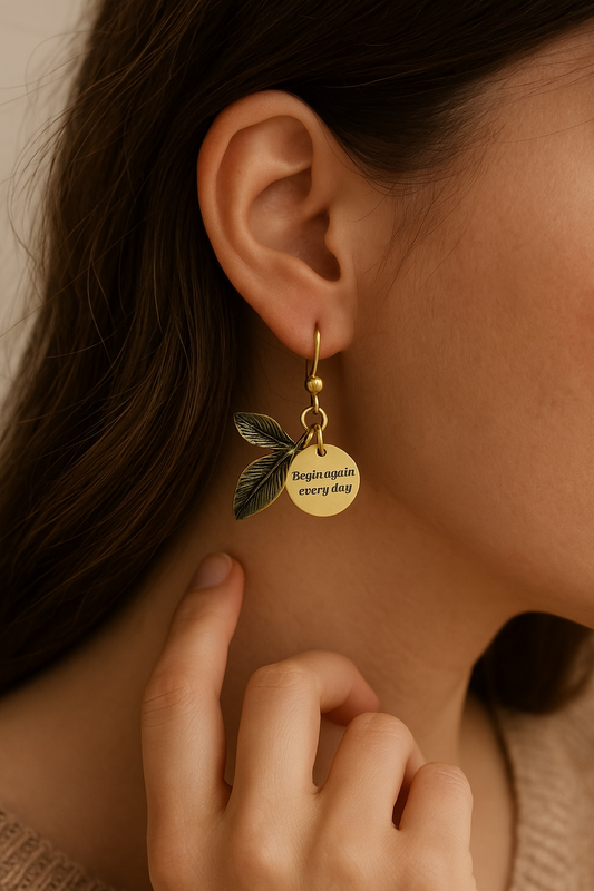 A close-up of a woman wearing an antique bronze and gold leaf-shaped earring. Attached to the earring is a small charm tag that reads, 'Begin again every day.'