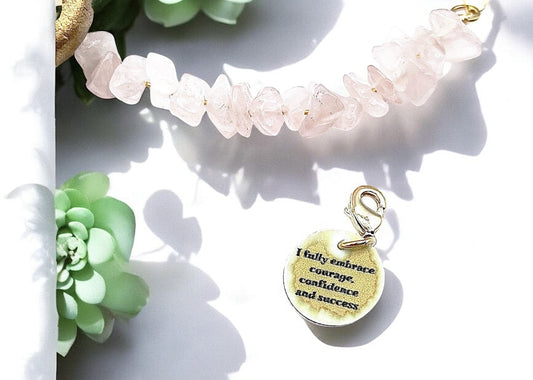 A gold rose quartz bracelet nestled among green succulents on a white background, featuring a charm inscribed: 'I fully embrace courage, confidence and success', blending natural elegance with soft, feminine tones.