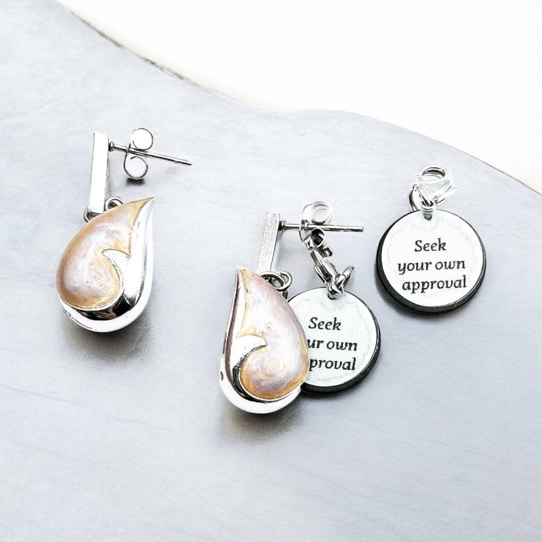 Silver nautilus shell earrings with charm tags that read 'Seek your own approval', beautifully arranged on a smooth grey surface - elegant, ocean-inspired, and uplifting.