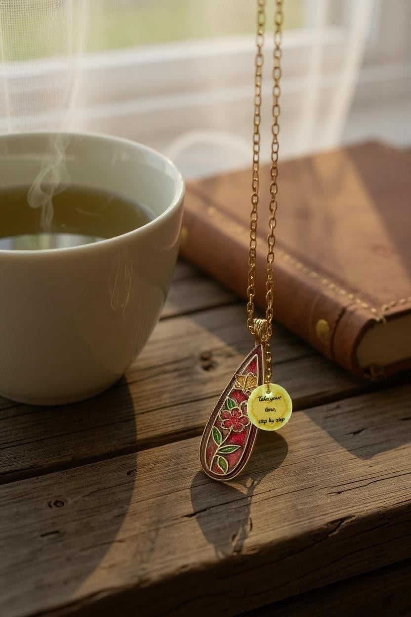 Floral enamel pendant necklace on a gold chain with a charm that reads 'Take your time, step by step', displayed on a wooden table between a cup of herbal tea and a leather-bound book.