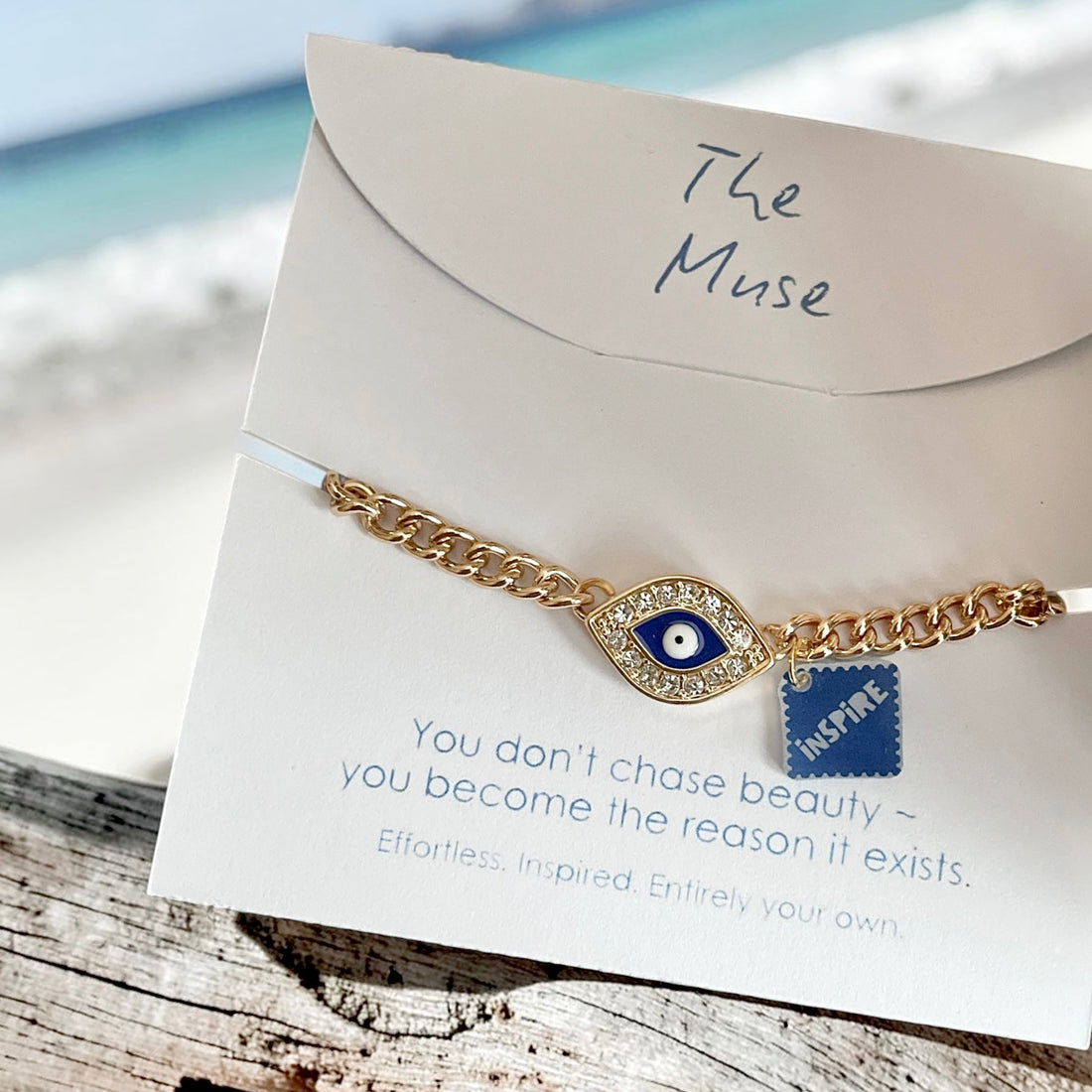 Gold evil eye bracelet with an inspire charm displayed on a white jewellery card against driftwood, with a soft-focus beach and sea in the background.