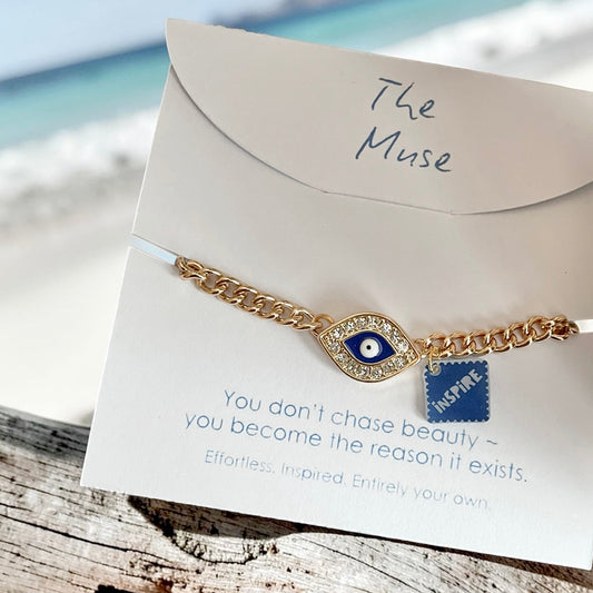 Gold evil eye bracelet with an inspire charm displayed on a white jewellery card against driftwood, with a soft-focus beach and sea in the background.
