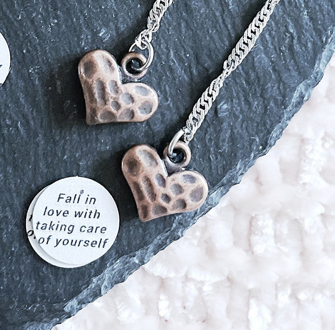 Bronze speckled heart pendant earrings on a slate tile, with a tag reading 'Fall in love with taking care of yourself'. 