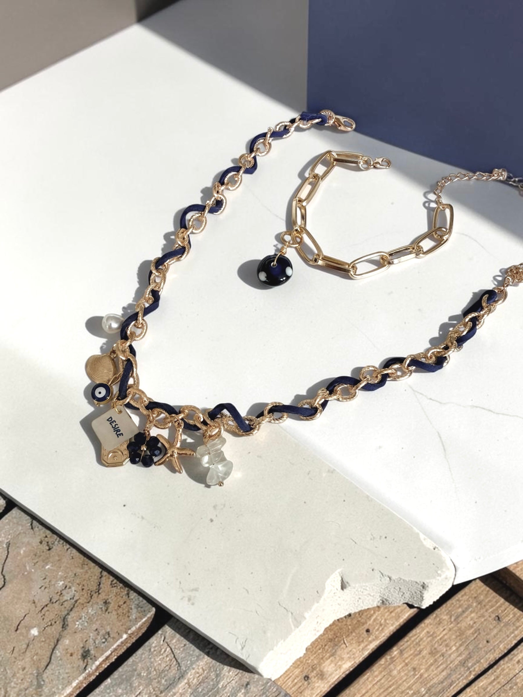 A gold-tone chain necklace and matching bracelet with dark blue beads and charms, displayed on a white stone slab outdoors.