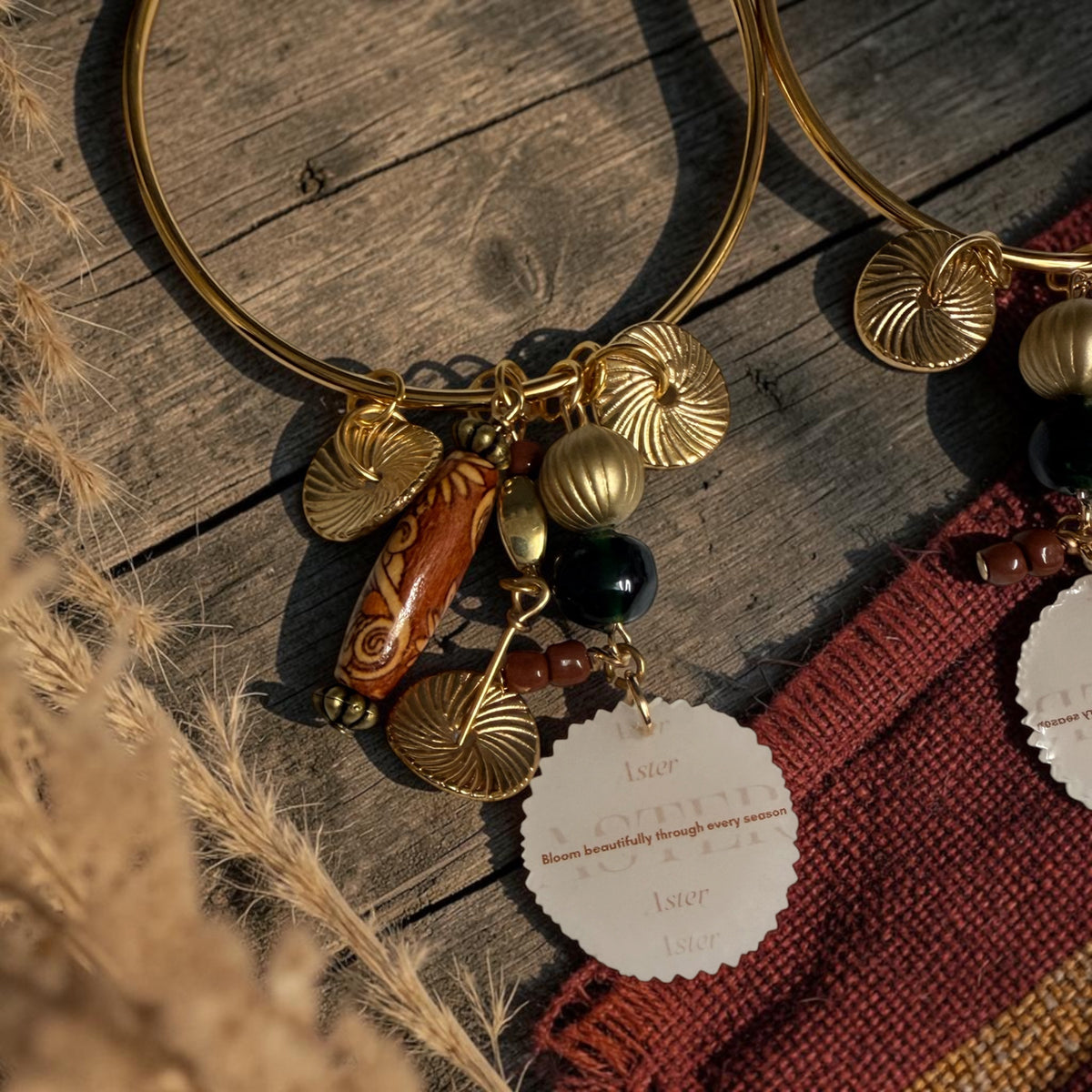 Gold-toned earring hoops with shell and bead charms, displayed on a rustic wooden surface with dried grass and a red textile.
