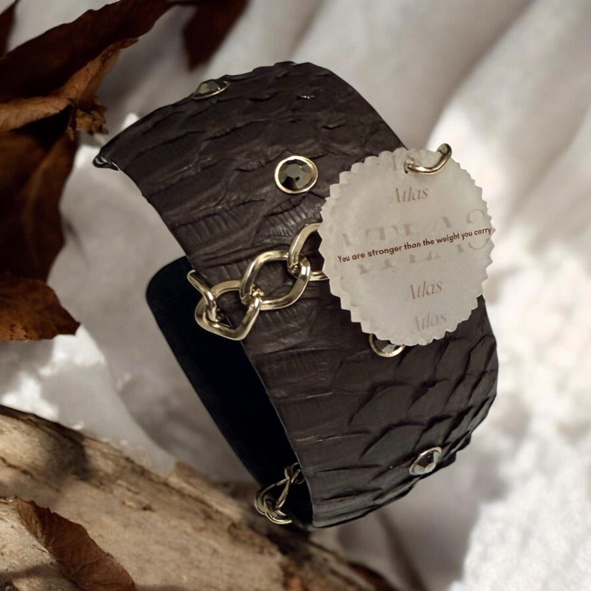 A dark brown textured leather cuff bracelet with gold-tone chain links and metallic embellishments, featuring a decorative reminder tag that reads 'You are stronger than the weight you carry'. The bracelet is styled on a piece of wood with dried leaves and white fabric.