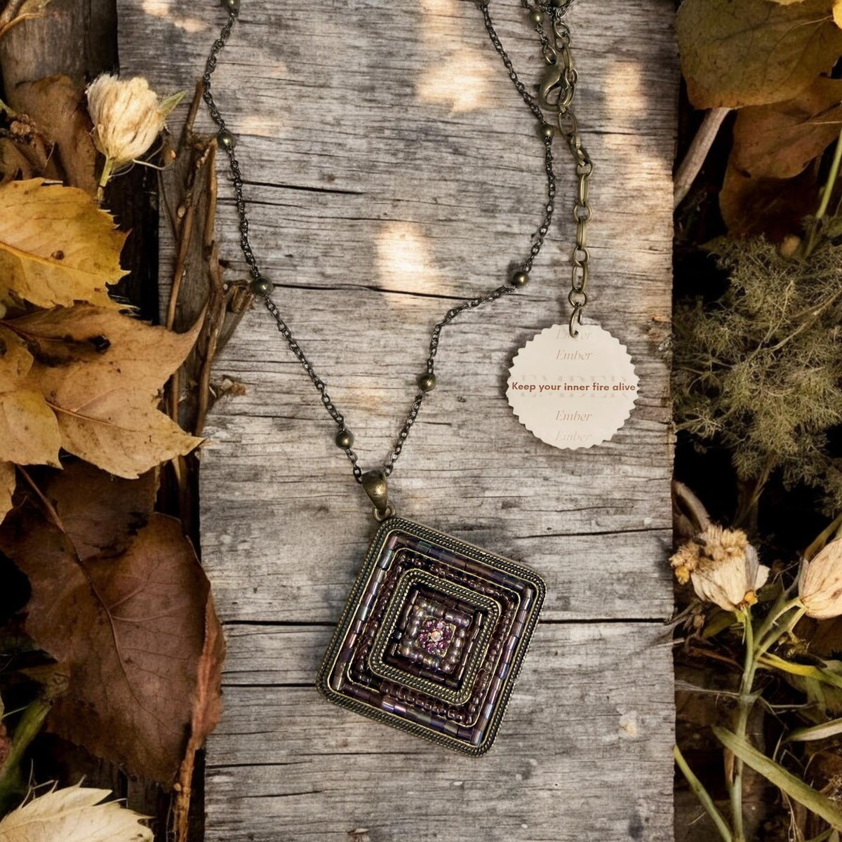 A geometric pendant necklace with layered metallic and purple-toned seed bead detailing, displayed on a weathered wooden surface with dried leaves and flowers. A small hanging tag reads 'keep your inner fire alive'.