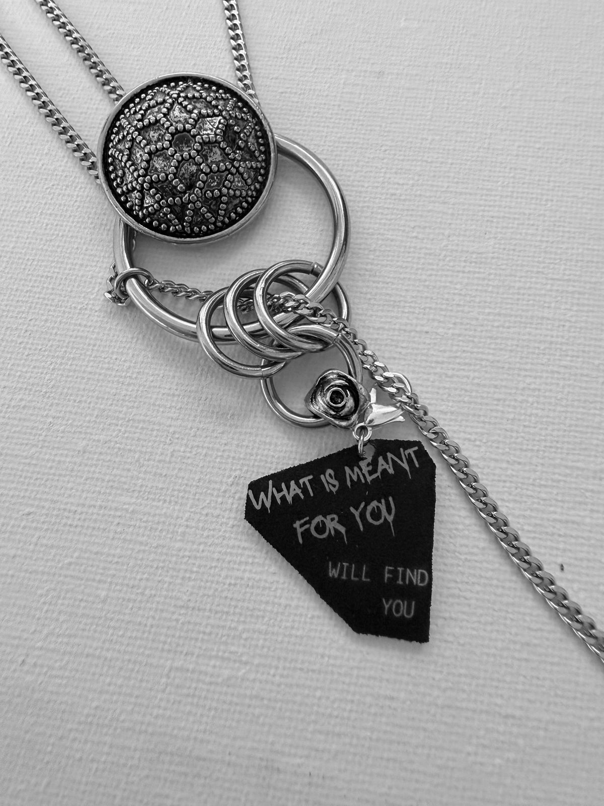 A silver-toned layered necklace with a textured round pendant, a large O-ring, and a dangling gothic style charm tag engraved with the words 'WHAT IS MEANT FOR YOU WILL FIND YOU' on a light fabric background.