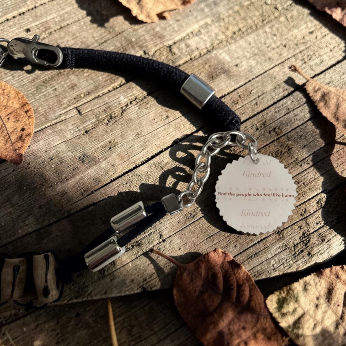 A black woven bracelet with silver metal accents, a lobster clasp, and a round white engraved charm printed with the phrase Find the people who feel like home', displayed on a rustic wooden surface with scattered dry leaves.