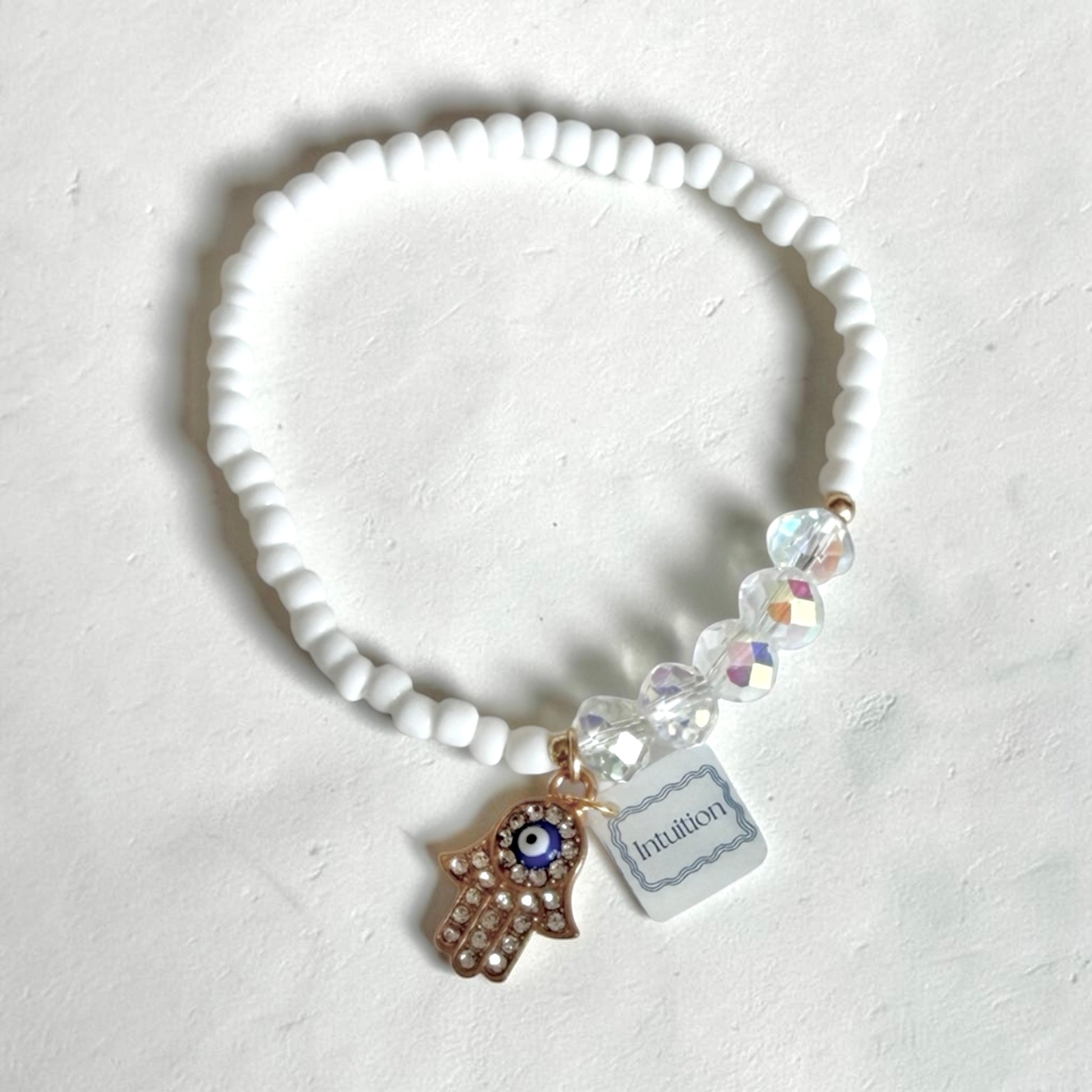 A white beaded stretch bracelet with a  gold Hamsa hand charm featuring a blue and white evil eye, clear faceted crystal beads, and a small white tag that reads 'Intuition'.