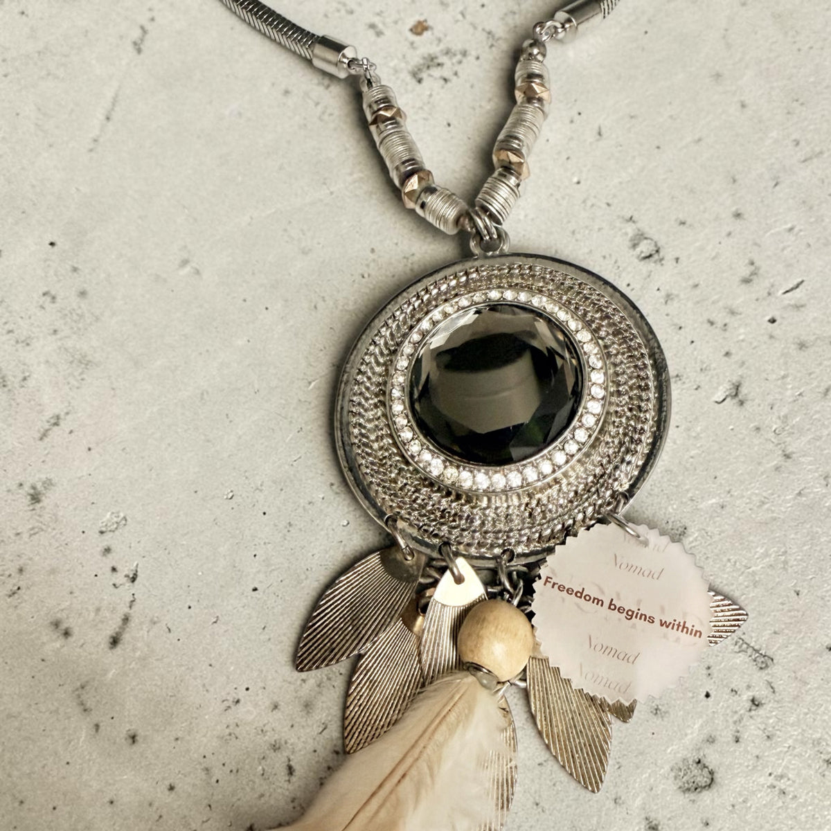 A bohemian-style silver pendant necklace featuring a large metallic bronze gemstone, textured silver rings, feather charms, and a cream feather. A reminder tag reading 'Freedom begins within' is attached.