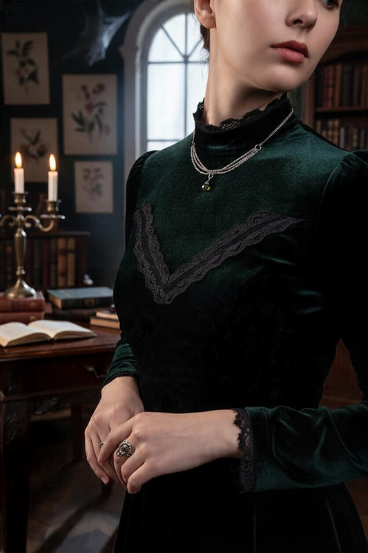 A lady is photographed wearing a green velvet dress and a silver-toned chain crystal pendant necklace. There is a desk with an open book, candelabra, floral wall pictures and an arch window in the background.