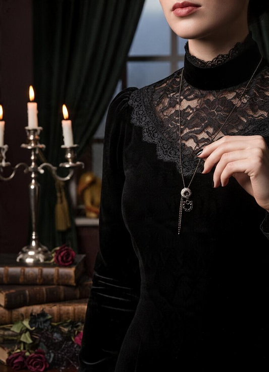 A close-up of a model wearing a black velvet Victorian-style dress with a lace mock neck and a gunmetal pendant necklace. The background is a dark, atmospheric room with lit candles, books, and dark drapery.