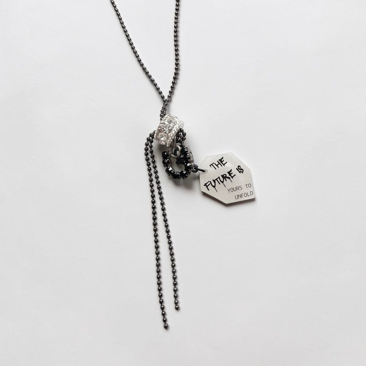 A gunmetal ball chain necklace with a horseshoe pendant, a cubic zirconia encrusted bead and a tag that reads 'THE FUTURE IS YOURS TO UNFOLD', photographed on a plain white background.