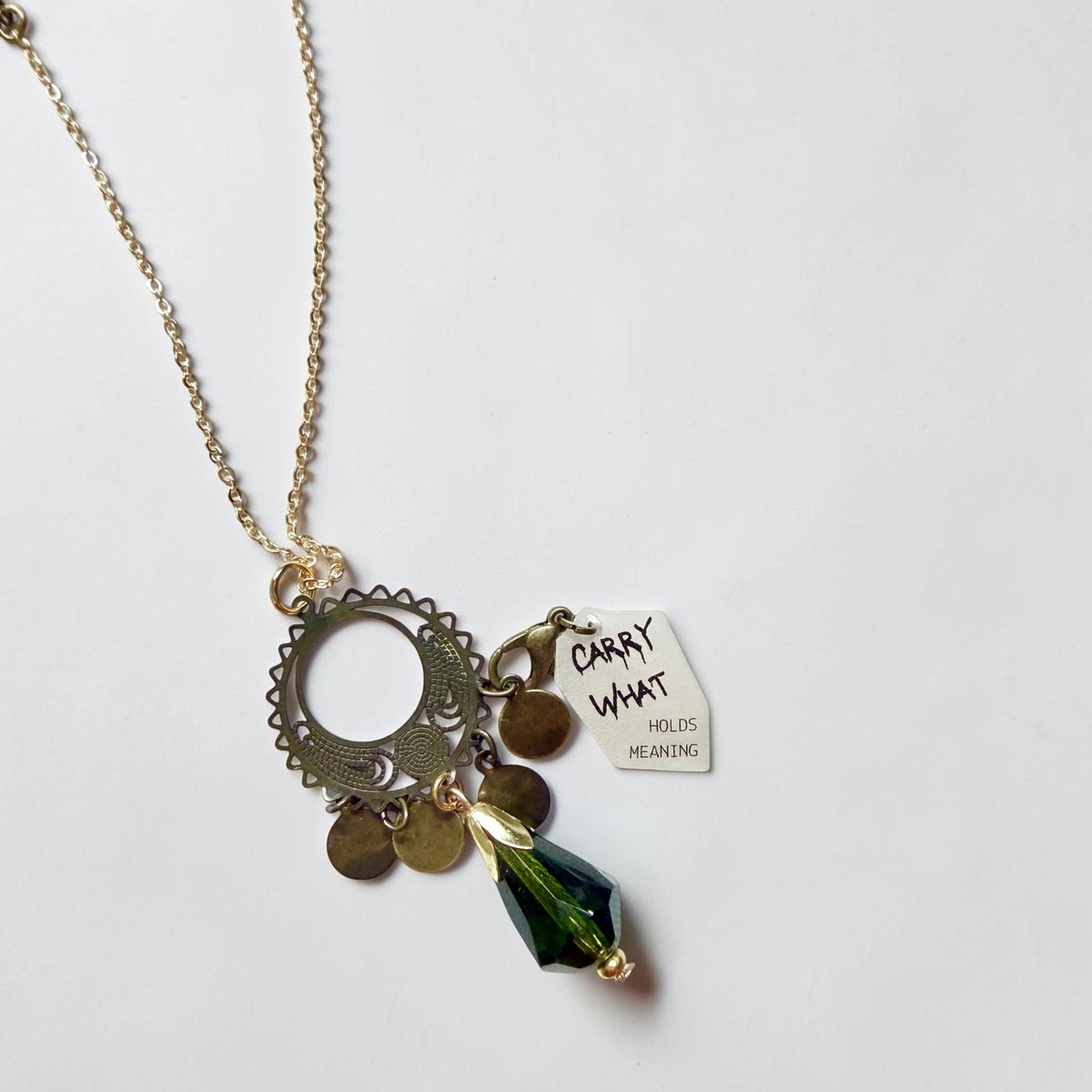 A gold-toned chain necklace with a circular metal pendant, small round charms, a white tag reading 'CARRY WHAT HOLDS MEANING' in black lettering, and a dark green faceted bead charm, photographed on a plain light background.