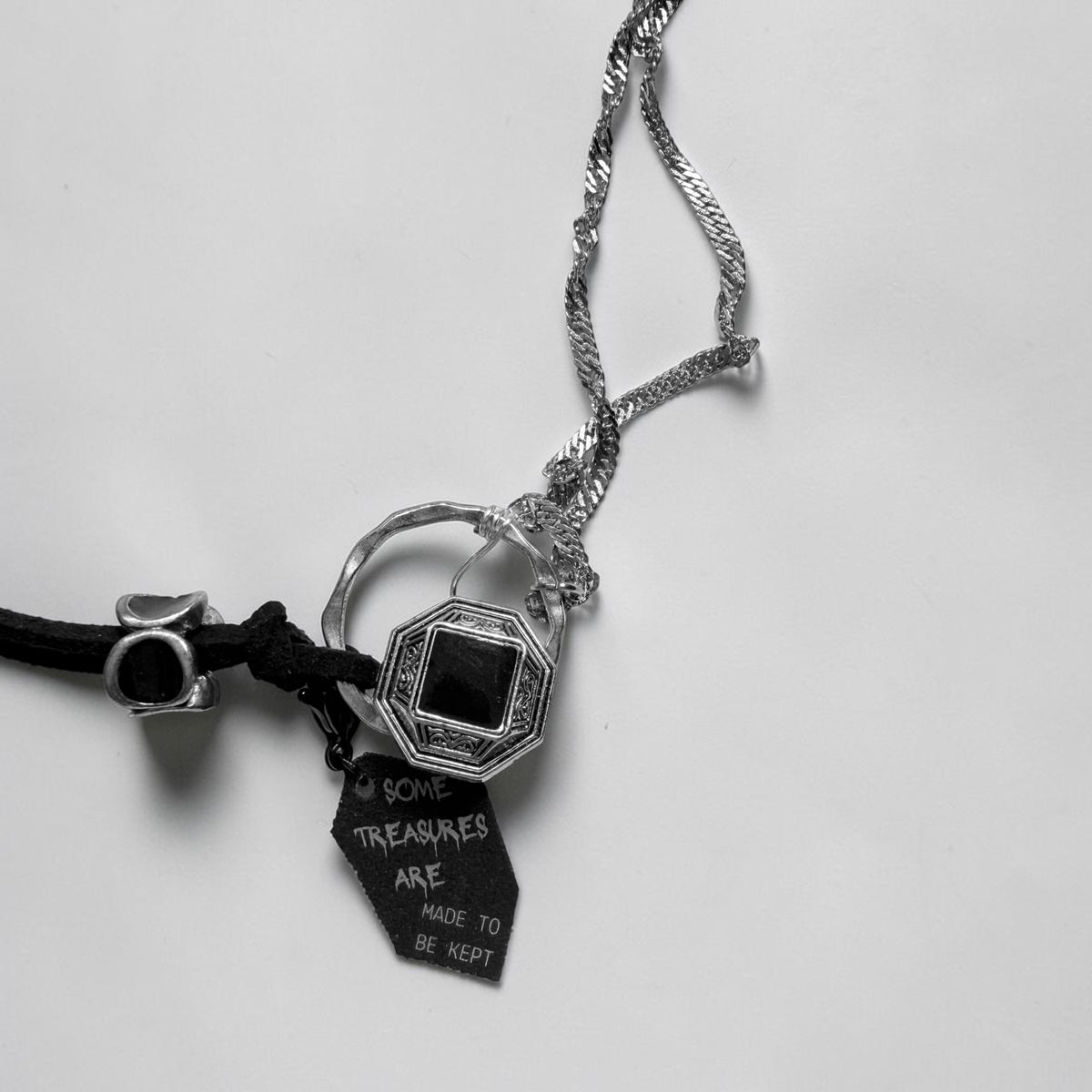 A gothic-style pendant necklace featuring a silver-toned octagonal charm with a black centre, black suede cord, stainless steel chains and a dangling black tag that reads 'SOME TREASURES ARE MADE TO BE KEPT'.