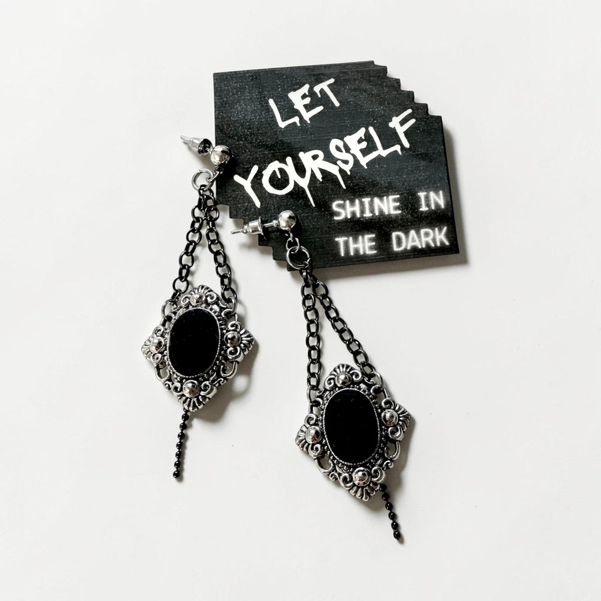 A pair of gothic-style dangle earrings with ornate silver filigree frames and black oval centres, hanging from black chains. They are displayed against a white background next to a black card with the text 'LET YOURSELF SHINE IN THE DARK'.