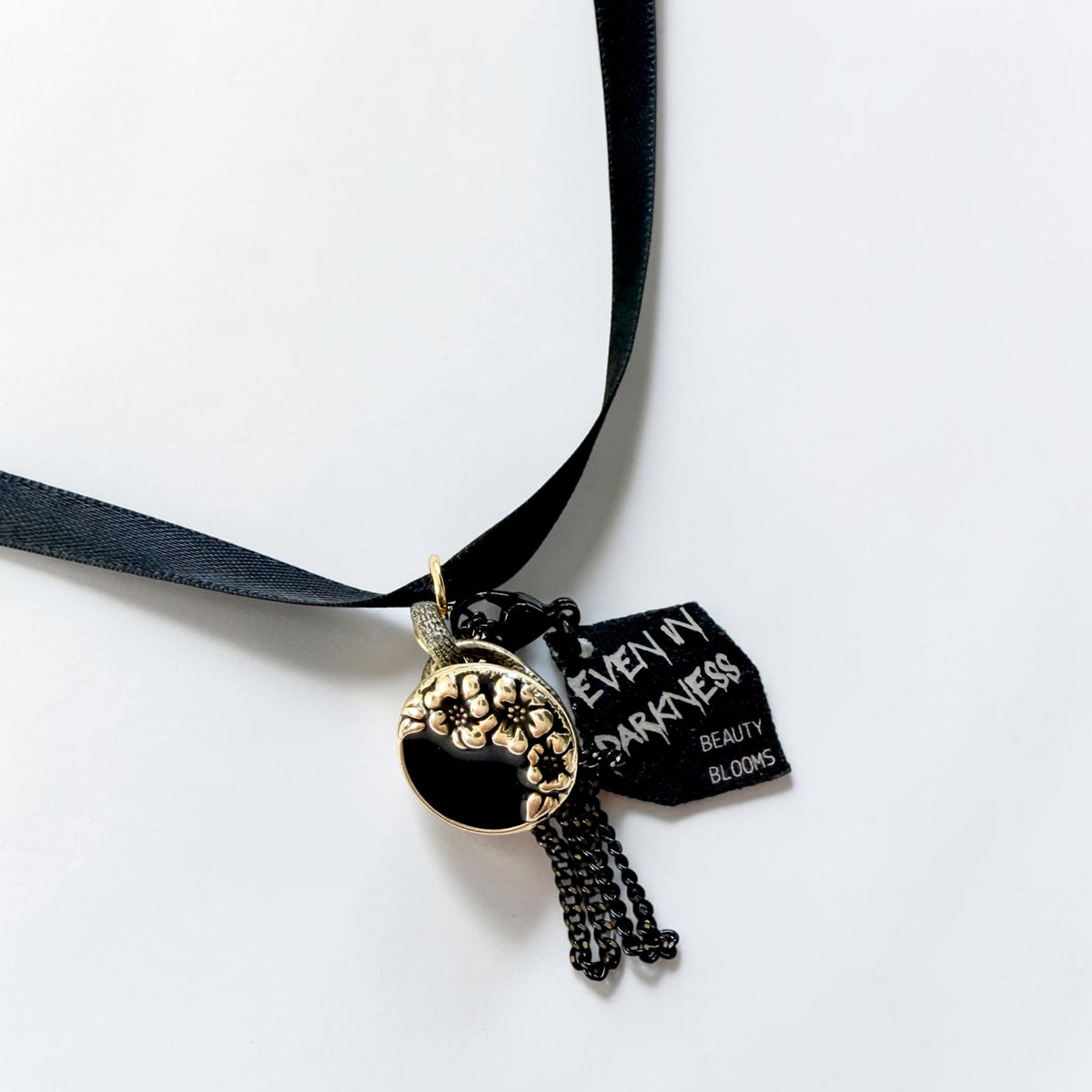 A gothic-style black ribbon choker necklace featuring a black and gold enamel pendant with a floral design, a black beaded tassel chain, and a black tag that reads 'EVEN IN DARKNESS BEAUTY BLOOMS'.