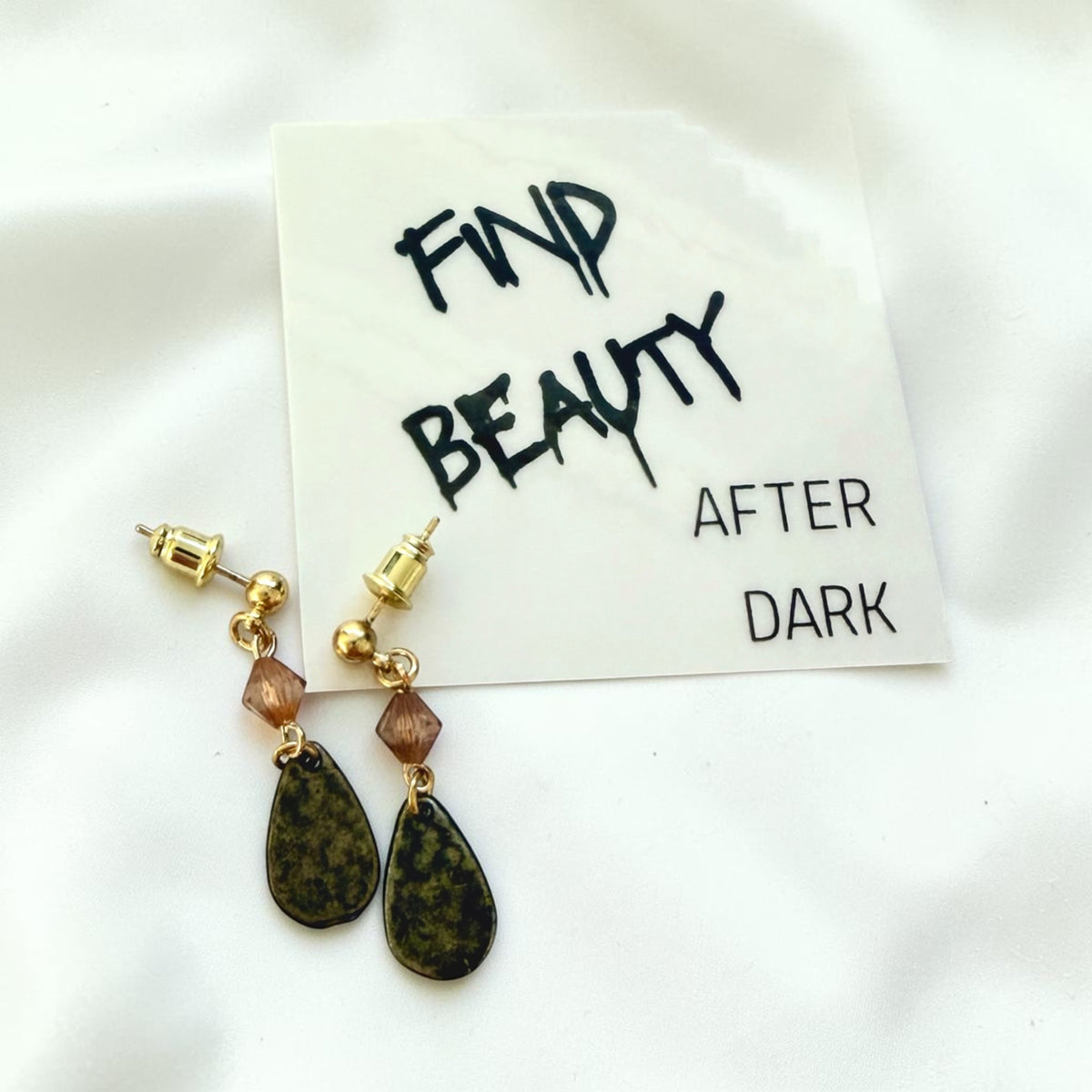 A pair of gold-tone dangle earrings with faceted brown beads and antique bronze teardrop pendants, displayed on a white card that reads 'FIND BEAUTY AFTER DARK' on a white fabric background.
