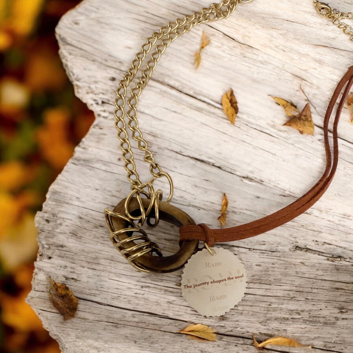 A close-up of a necklace laid on weathered white wood with scattered dry leaves. The necklace has a gold-toned chain, an oval wood-effect pendant with oval link rings attached, a dark tan suede cord, and a small round tag with engraved text.