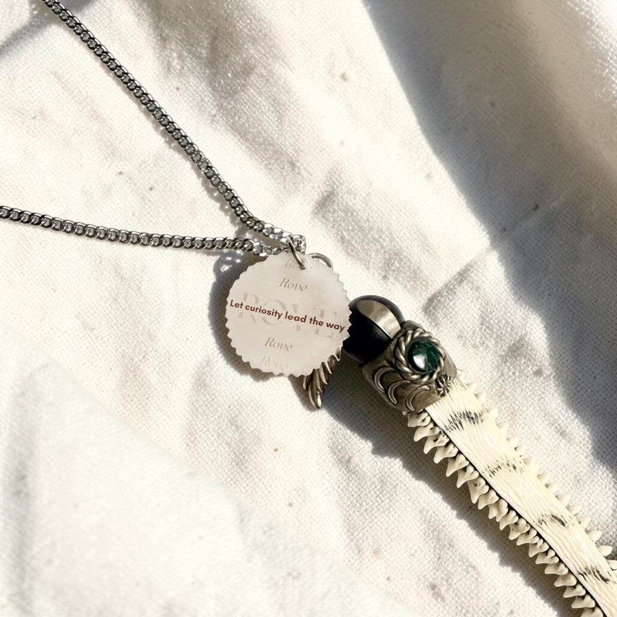 A silver-toned chain necklace with a sawtooth tooth charm and a round engraved tag, displayed on a textured off-white fabric under direct sunlight.
