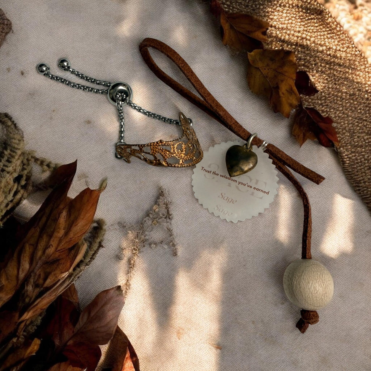 A bohemian-style jewellery set featuring a copper tone filigree slider ring, and a hanging suede keepsake reminder, styled on a textured beige fabric with dried autumn leaves.
