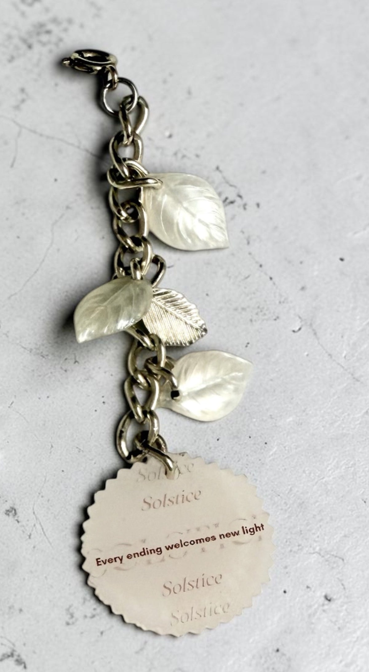 A silver-toned metal charm hanging keepsake with three leaf charms and a round tag that reads 'Every ending welcomes new light', photographed on a textured light grey surface.