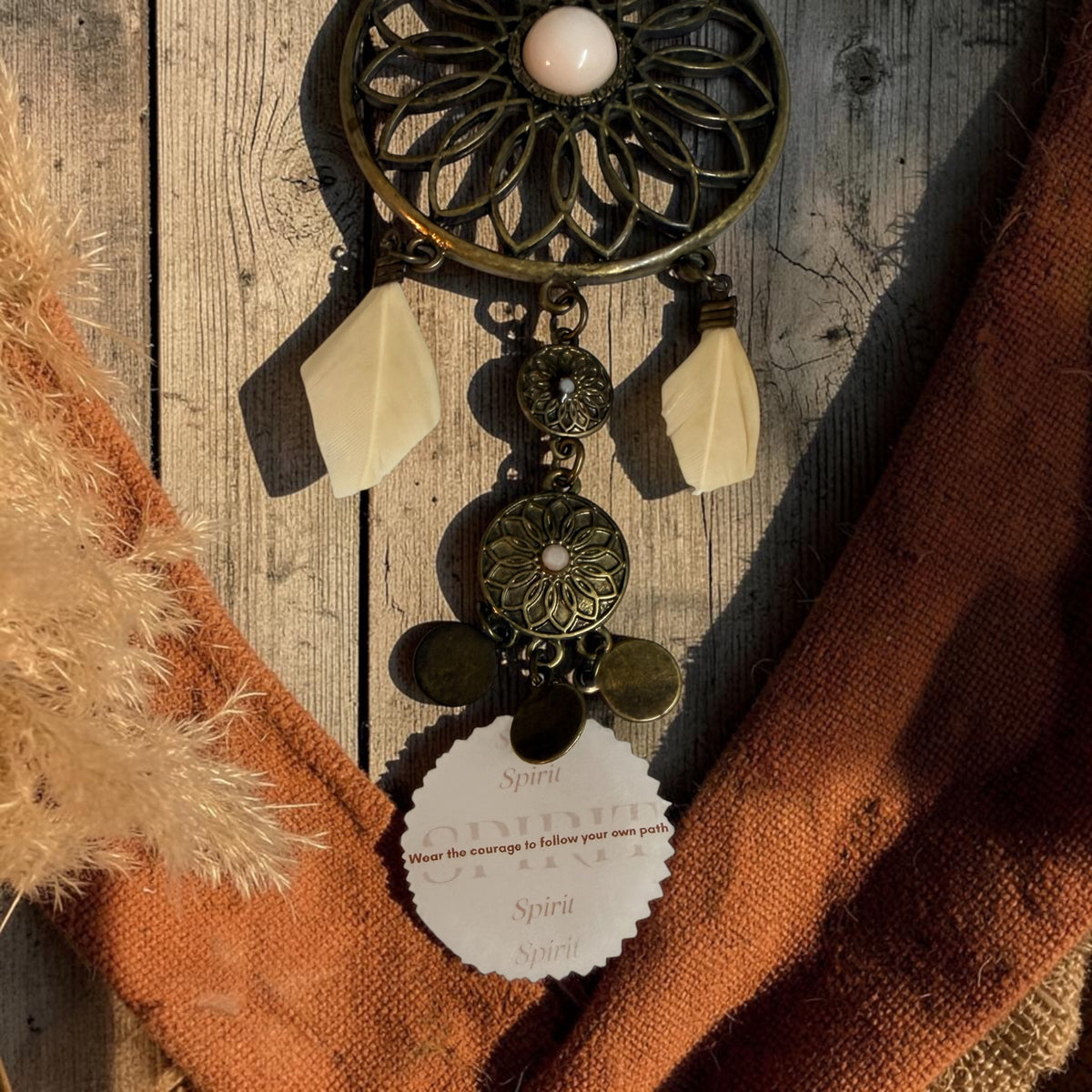 A bohemian-style dreamcatcher necklace with an antique bronze circular mandala-style pendant, blush pink glass beads, coin and feather charms, and a circular reminder tag that reads, 'Wear the courage to follow your own path', displayed on a rustic wooden surface with an orange scarf and dried grass as props.