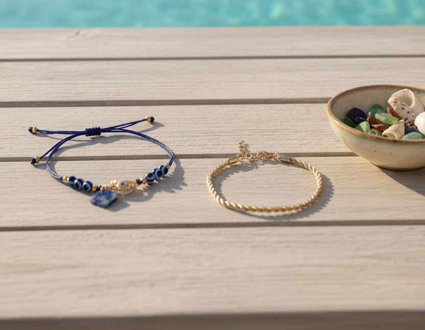 Two delicate anklets displayed on a light wood surface with a bowl of seashells nearby and a blurred blue water background. One anklet is navy blue cord with small beads and a blue stone charm, and the other is a gold-toned twisted rope chain anklet.