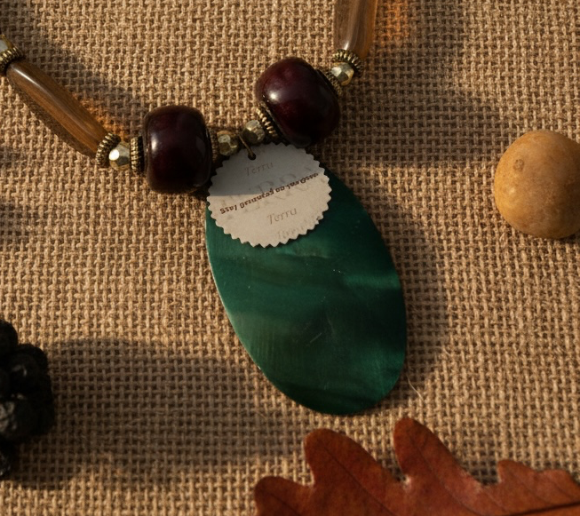 A handmade beaded necklace featuring a large oval green pendant, dark reddish-brown wooden beads, translucent brown beads, and a circular reminder tag reading 'Stay grounded as you grow', displayed on a burlap background with decorative leaves.