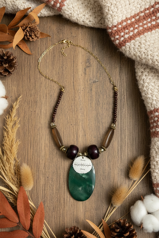 A beaded necklace with chain, featuring a large oval green pendant, dark reddish-brown wooden beads, translucent brown beads, and a circular reminder tag reading 'Stay grounded as you grow', displayed on a wooden background with a cosy blanket and foliage surround.