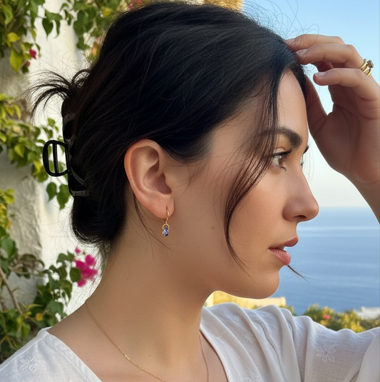 A woman in a white embroidered top wearing a delicate blue stone gold earring, photographed outdoors with a sea and wall in the background.