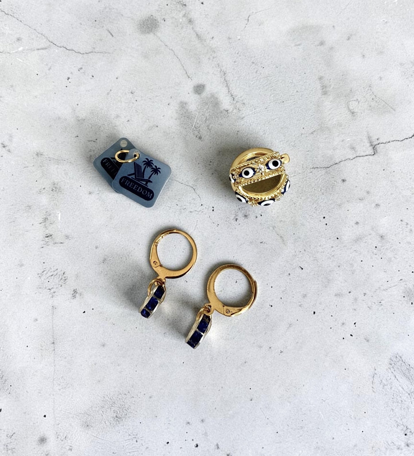 Two pairs of gold-toned earrings and word charms laid flat on a light grey stone-like surface: small blue square charm tags with black graphic and text labelled 'Freedom', a gold pair of huggie hoops with evil eye detailing, and a pair of gold hoop earrings with dark blue rectangular drops.