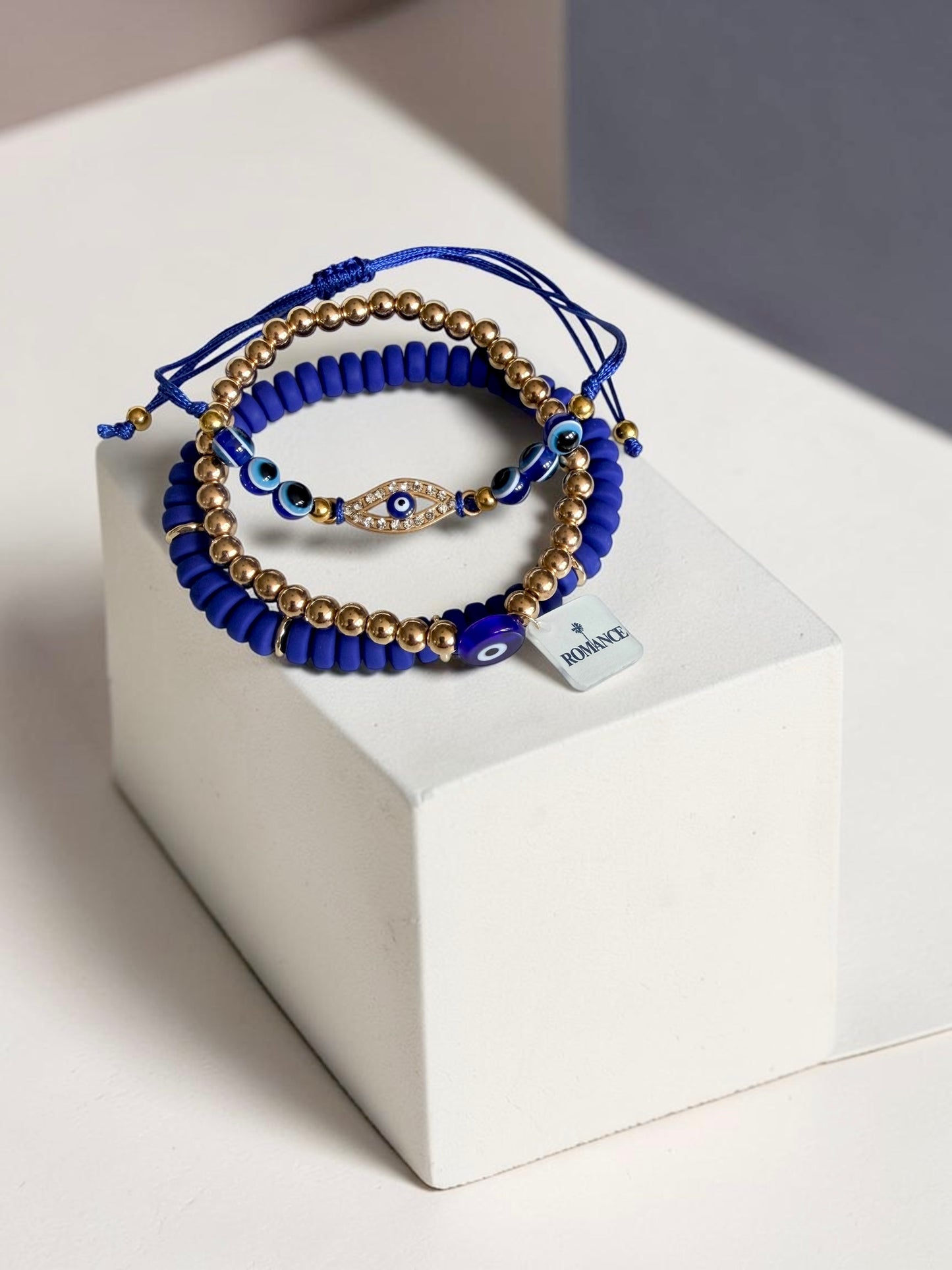 A stack of blue and gold beaded bracelets with evil eye charms, displayed on a white geometric block. One bracelet has a small square tag with the word 'Romance'.