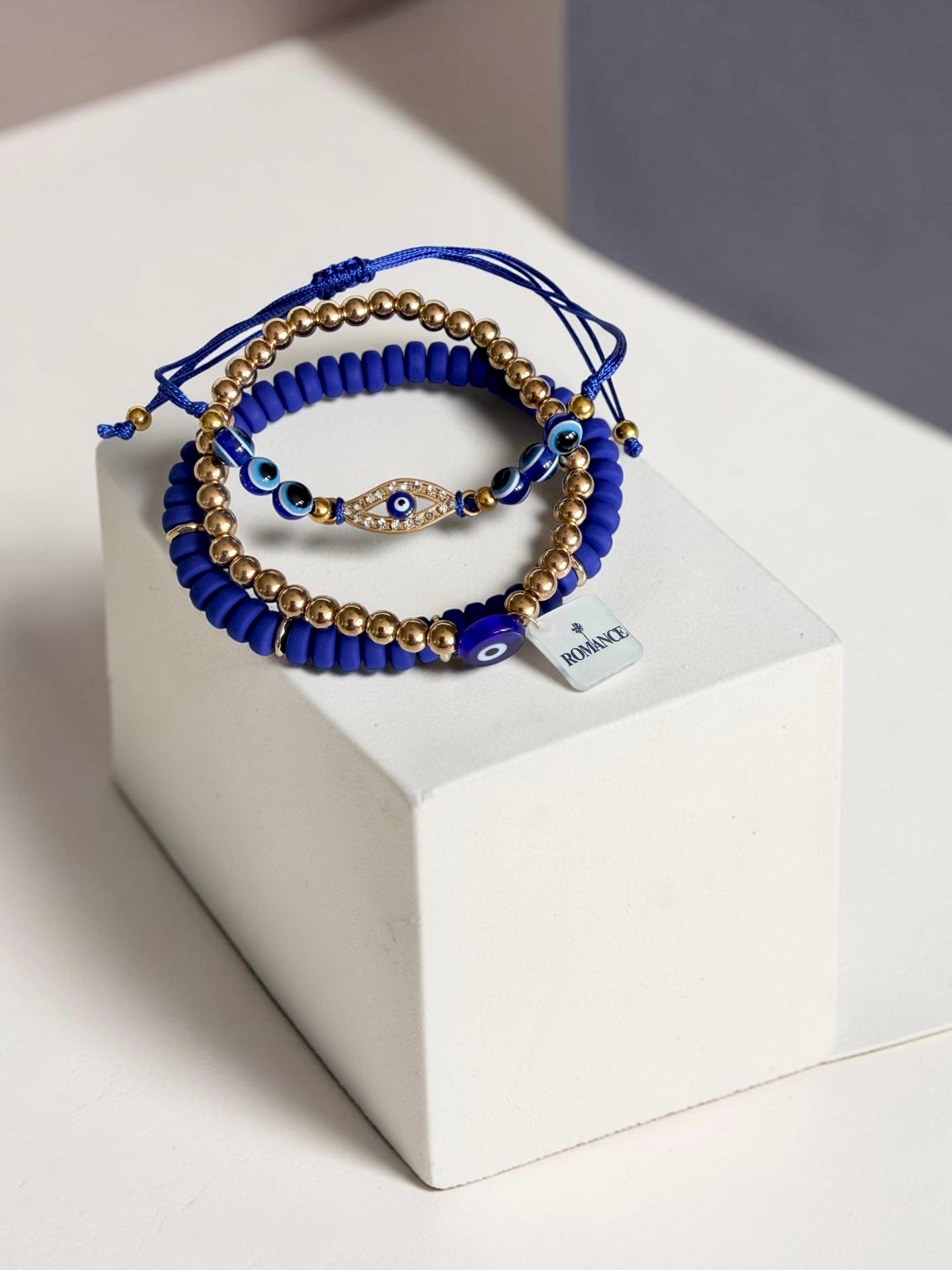 A stack of blue and gold beaded bracelets with evil eye charms, displayed on a white geometric block. One bracelet has a small square tag with the word 'Romance'.