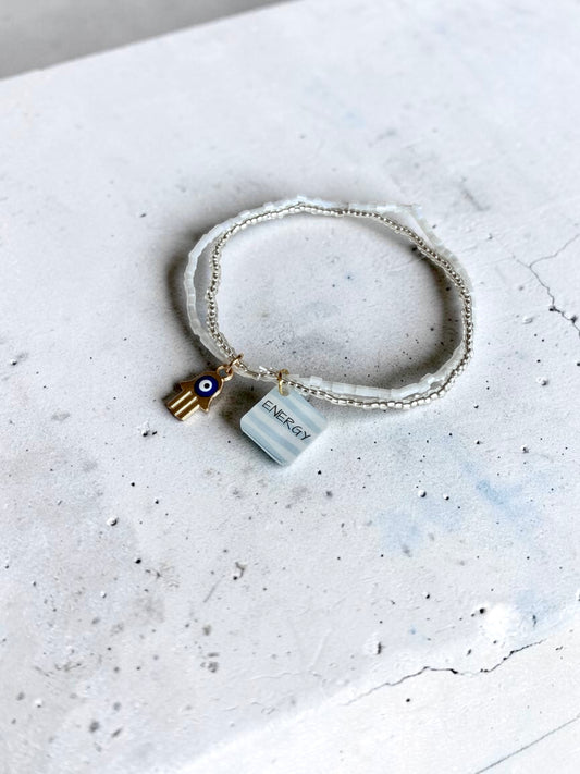 A delicate white beaded bracelet with a small gold Hamsa charm featuring a blue and white evil eye, and a white square  charm tag engraved with 'ENERGY', displayed on a textured light grey surface.