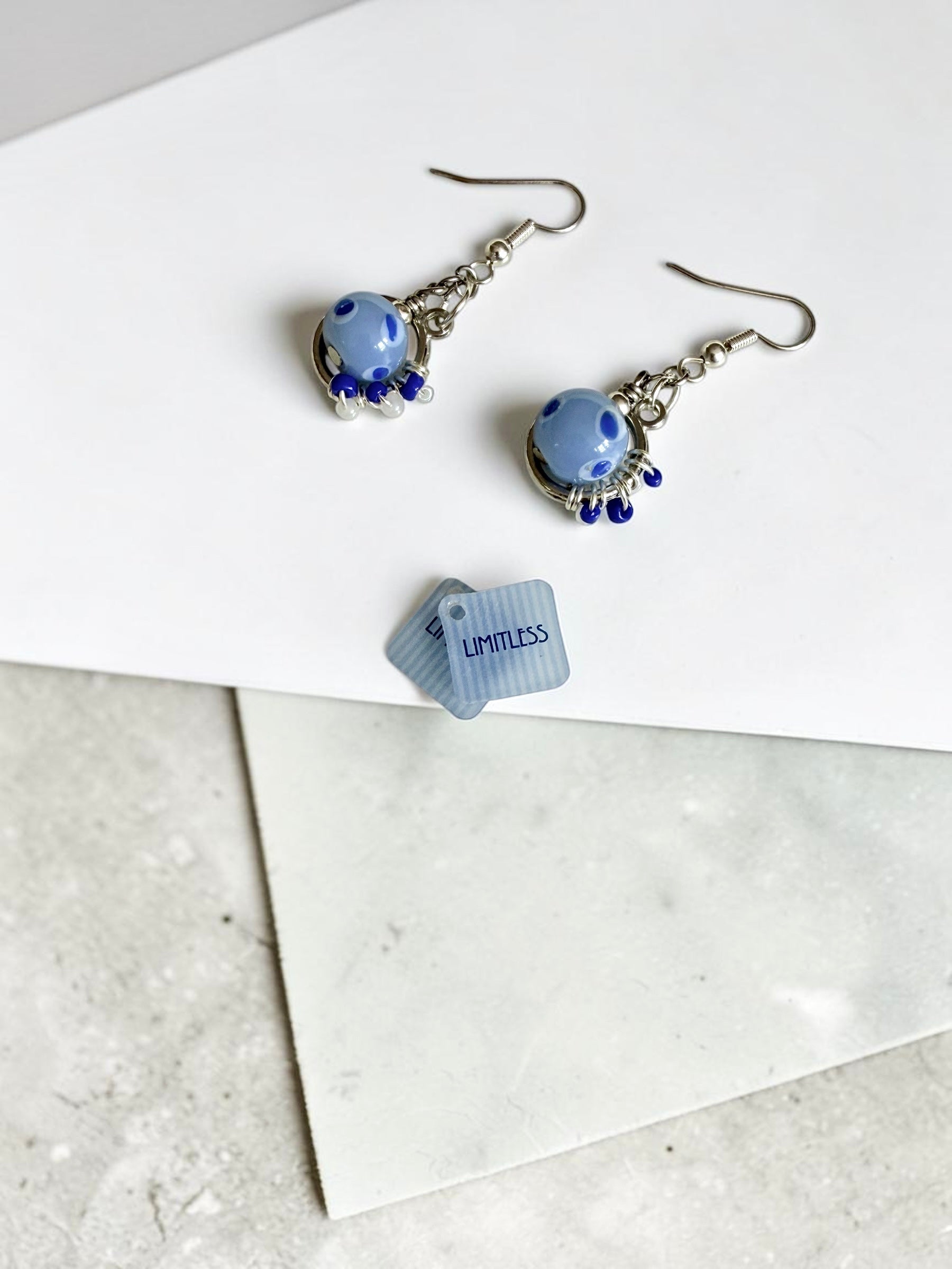 A pair of dangling earrings with light blue marbled beads, silver-toned hooks, and small dark blue bead drops, displayed on a white card over a light grey marble surface. A couple of small tags reading 'LIMITLESS' are also visible.